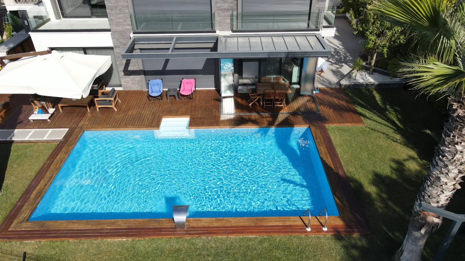 Villa bahçesinde ahşap deckli dikdörtgen prefabrik havuz, kenarında paslanmaz şelale ve merdiven