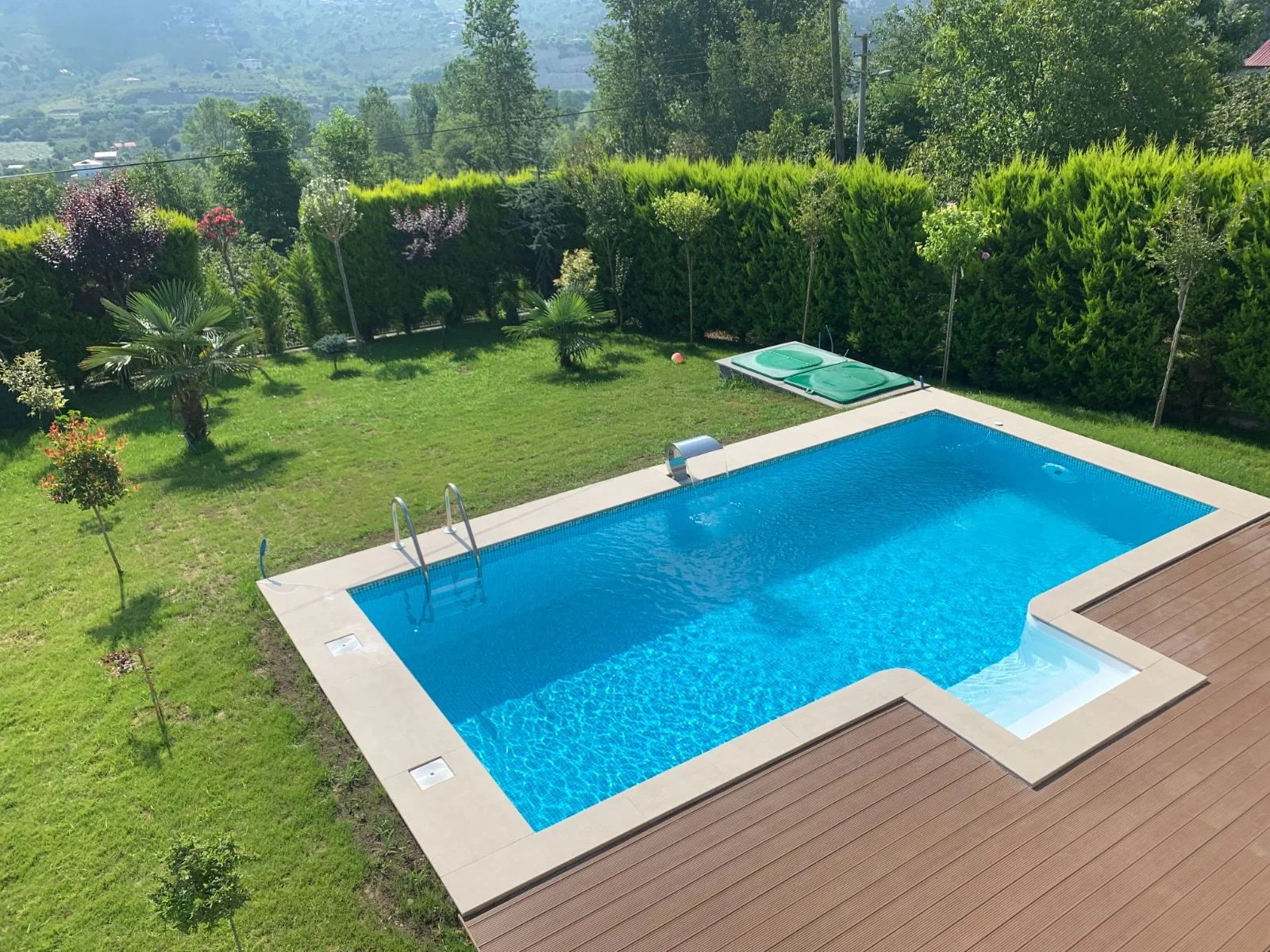 Yamaç bahçede kurulmuş L formlu panel havuz, geniş deck ve basamak bölgesiyle birlikte