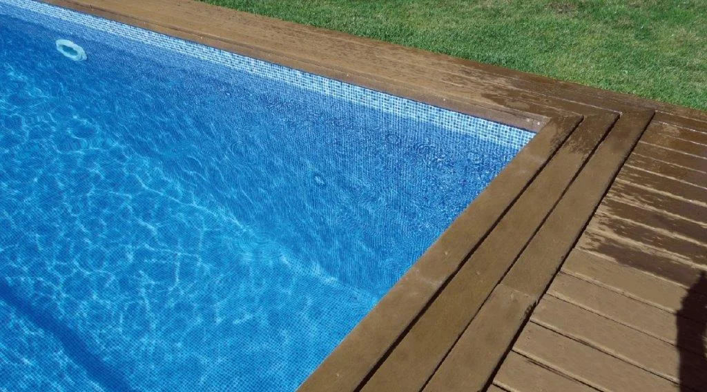 Ahşap deck ile çevrili havuzun köşesi — kenar, su yüzeyi ve çim geçişi bir arada